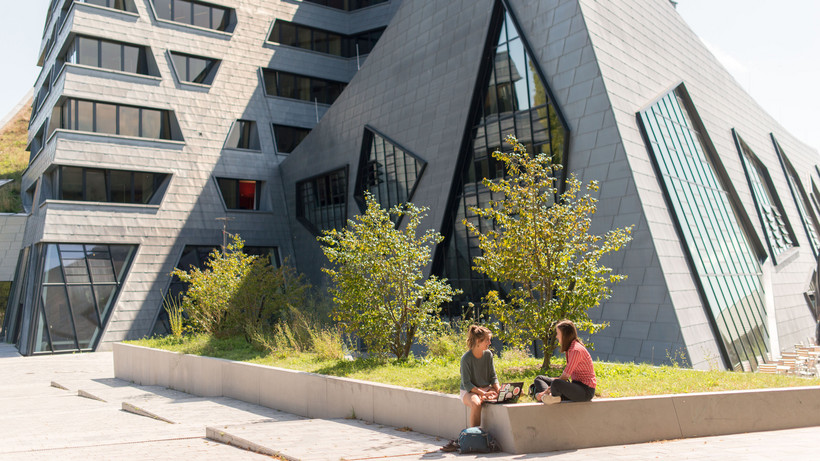 Two students sitting in the greenery in front of Leuphana's central building.